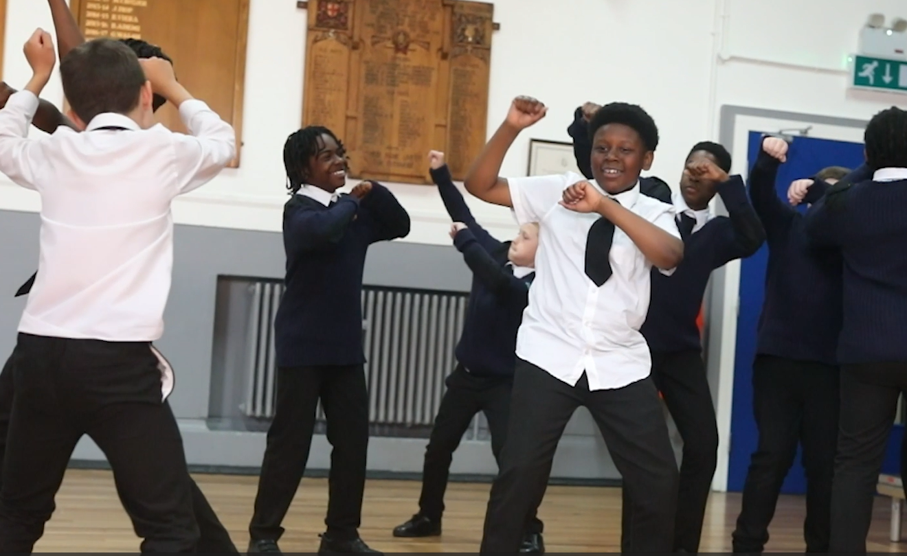 Children in school uniforms dance enthusiastically in a classroom setting; one child in a white shirt is prominent in the foreground, while others in blue sweaters join in the background.