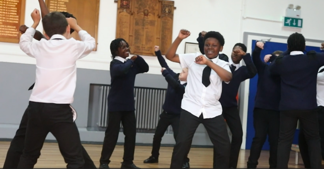 Children in school uniforms dance enthusiastically in a classroom setting; one child in a white shirt is prominent in the foreground, while others in blue sweaters join in the background.