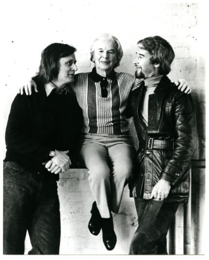 Three people, one seated with two others standing close, posing for a photo against a plain background. The seated person's arms are around the shoulders of the two standing individuals.