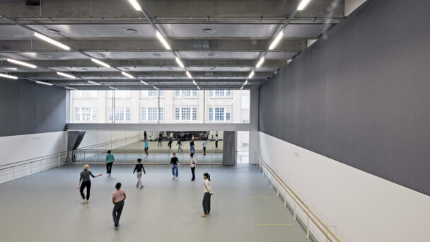 A spacious dance studio with high ceilings, large mirrors, and natural light from wall-length windows. Several people are practicing dance, dispersed throughout the room.