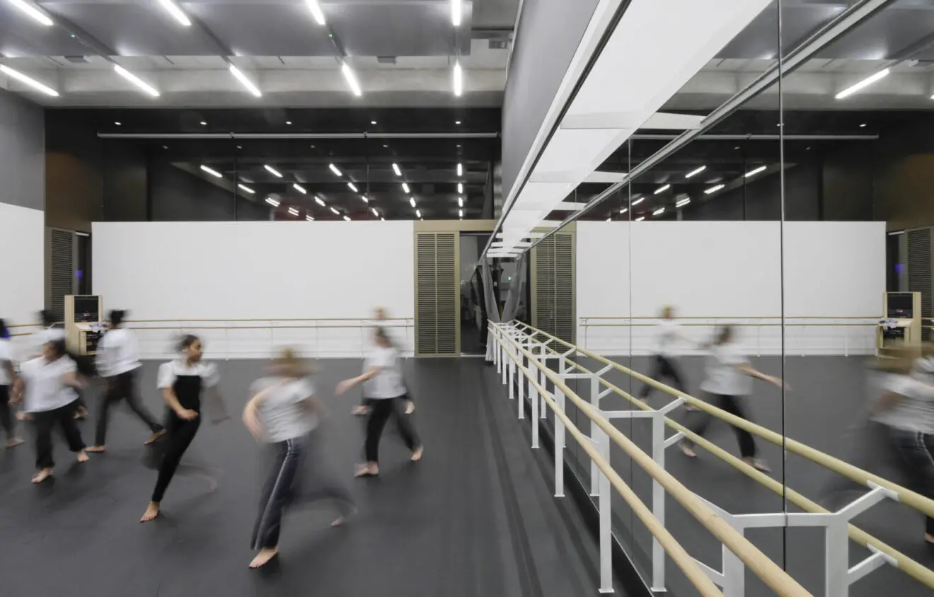 A group of dancers in motion during a rehearsal in a large, modern studio with floor-to-ceiling mirrors and ballet barres. The studio is well-lit with overhead lighting.