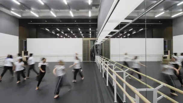 A group of dancers in motion during a rehearsal in a large, modern studio with floor-to-ceiling mirrors and ballet barres. The studio is well-lit with overhead lighting.