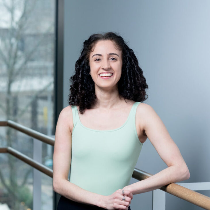 A person with curly hair in a green top smiles while leaning on a wooden handrail indoors, with a large window and trees visible in the background.
