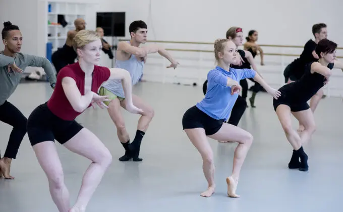 A group of dancers is practicing in a studio, performing synchronized movements with focused expressions. They are dressed in various workout clothes and appear to be concentrating on their routine.