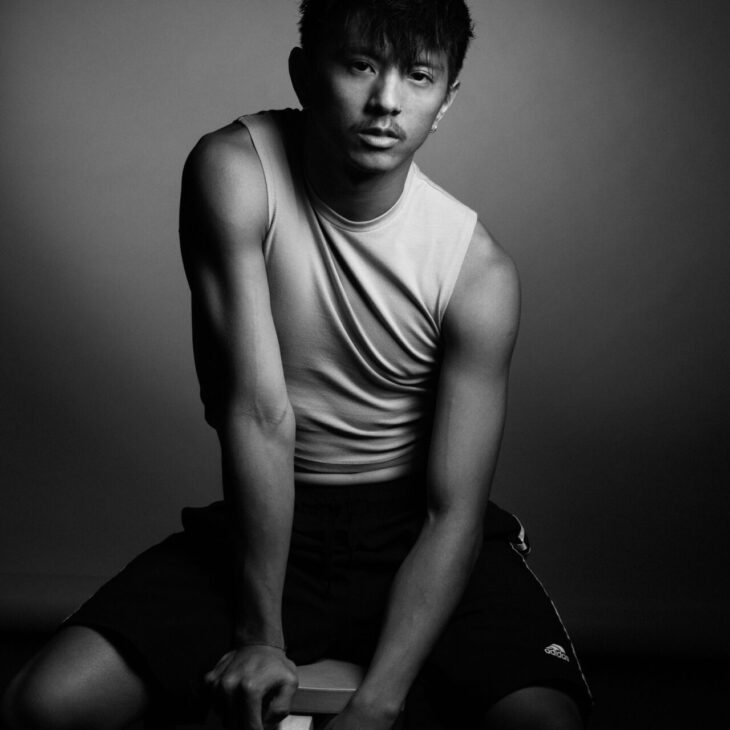 A black-and-white image of a person with short hair, seated on a stool, wearing a sleeveless shirt and shorts, looking directly at the camera.