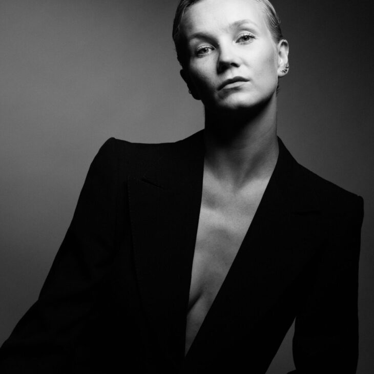 A black and white portrait of a person with short hair, wearing a dark blazer with a bare chest underneath, set against a plain background.