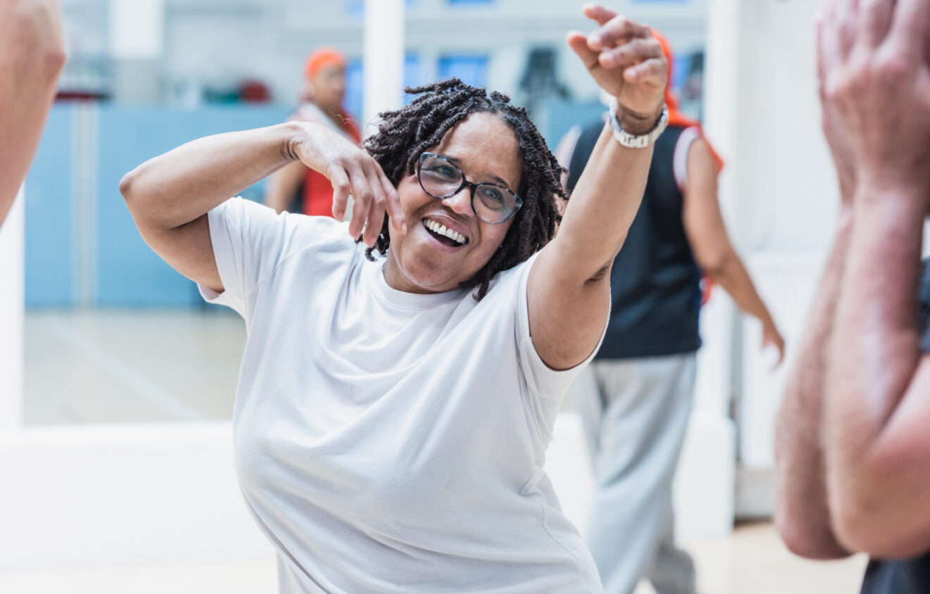 A woman with glasses and braided hair, wearing a white shirt, dances joyfully in a well-lit room with others.