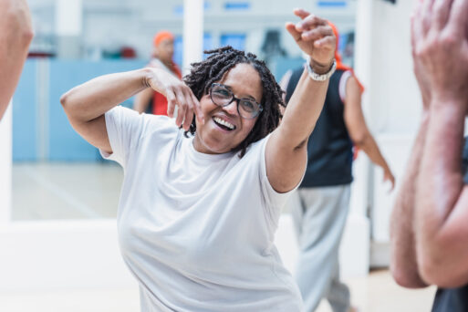 A woman with glasses and braided hair, wearing a white shirt, dances joyfully in a well-lit room with others.