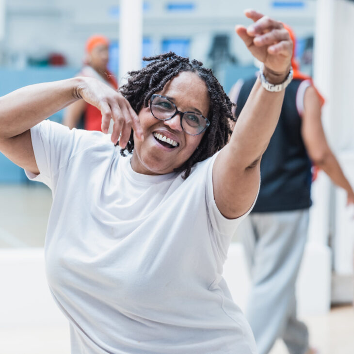 A woman with glasses and braided hair, wearing a white shirt, dances joyfully in a well-lit room with others.