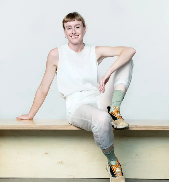 A person with short hair, dressed in white athletic wear, is sitting on a bench, resting one arm behind them and one foot on the bench, smiling at the camera.