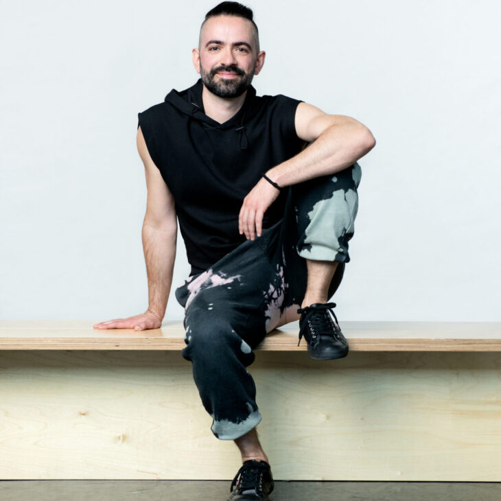 A man with a beard and mustache, wearing a sleeveless black hoodie and tie-dye pants, sits on a wooden bench with one leg up and his arm resting on his knee.