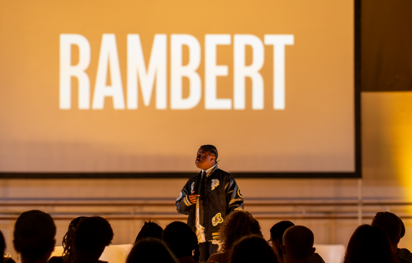 A person stands on stage, speaking to an audience with a large screen behind them displaying the word "RAMBERT.