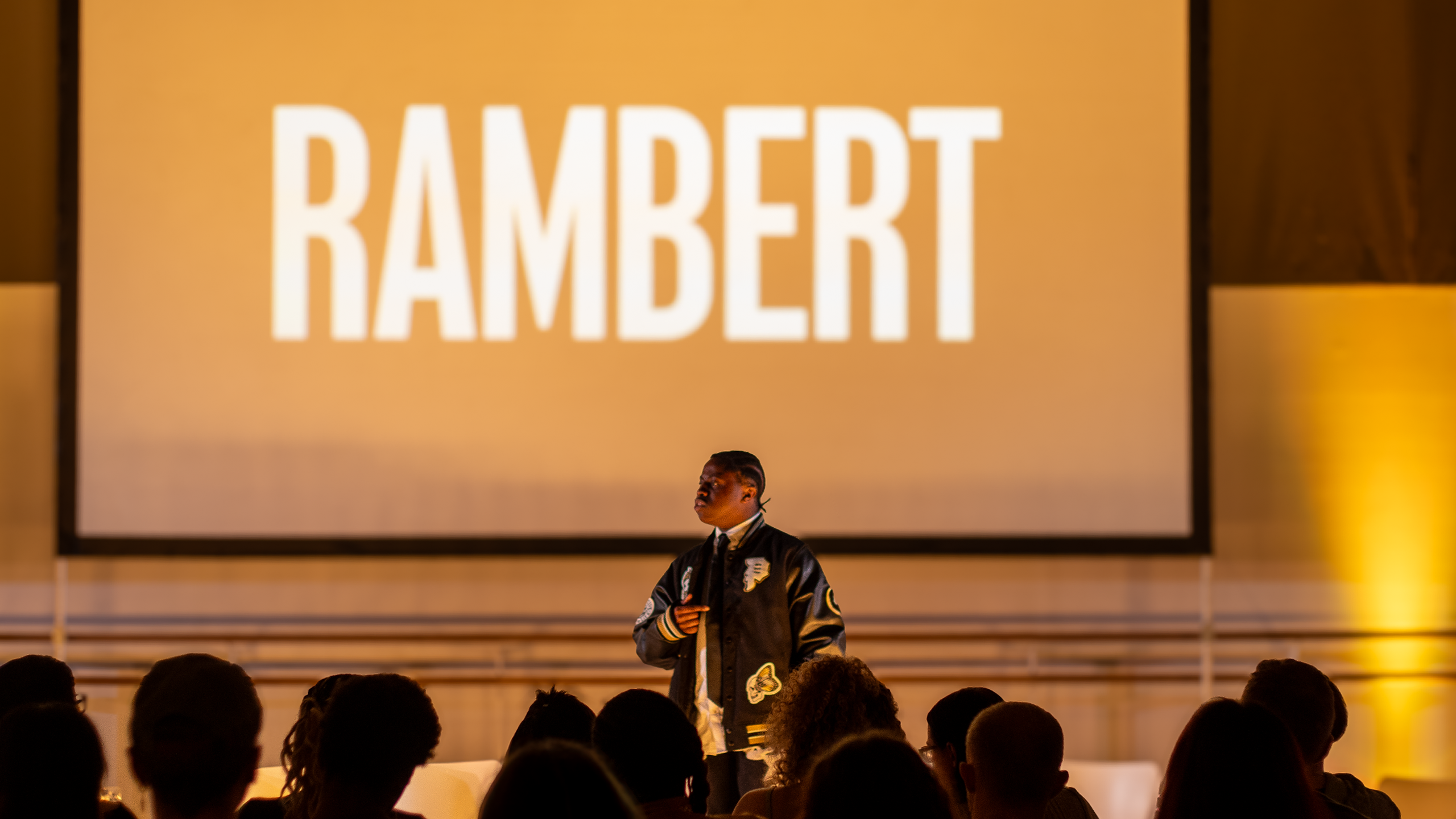 A person stands on stage, speaking to an audience with a large screen behind them displaying the word "RAMBERT.
