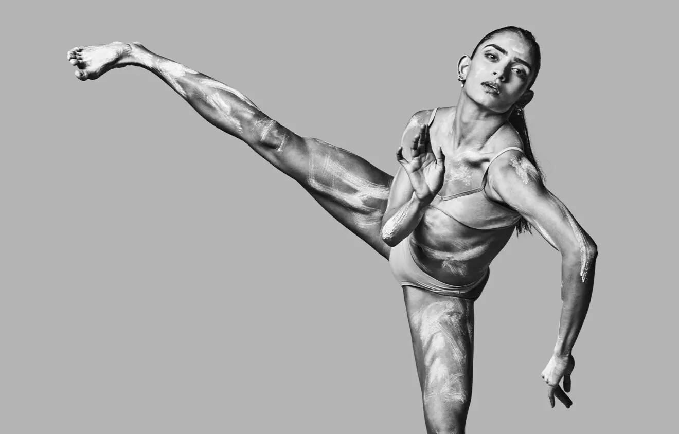 A female dancer performs a high kick, displaying flexibility and muscle definition, in a grayscale photograph.