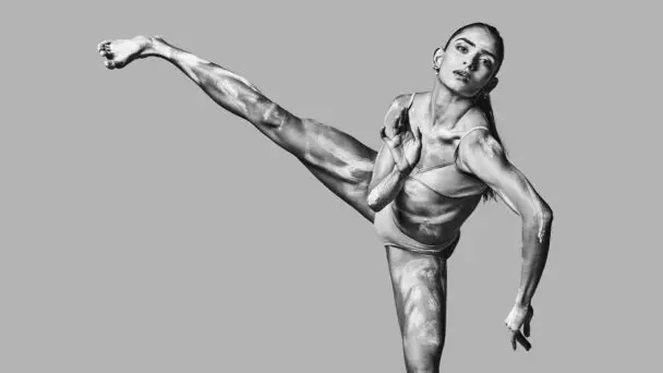 A female dancer performs a high kick, displaying flexibility and muscle definition, in a grayscale photograph.