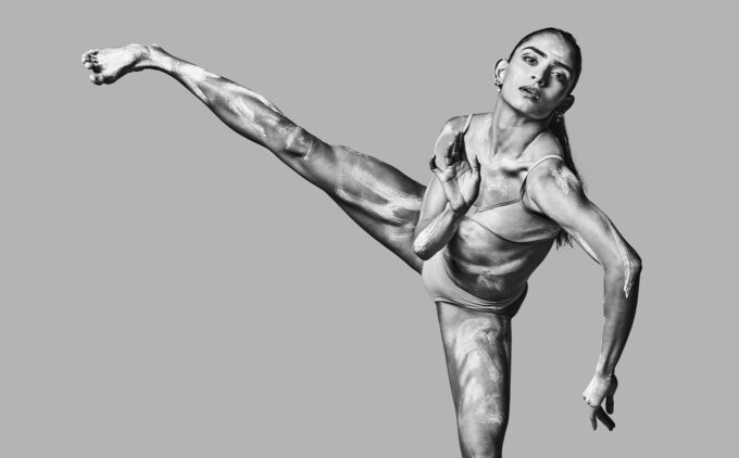 A female dancer performs a high kick, displaying flexibility and muscle definition, in a grayscale photograph.