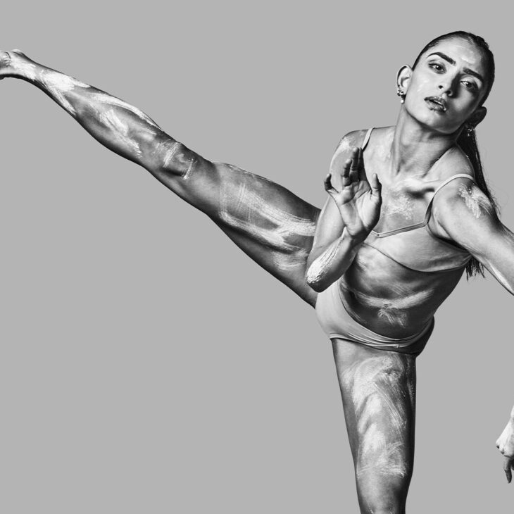 A female dancer performs a high kick, displaying flexibility and muscle definition, in a grayscale photograph.