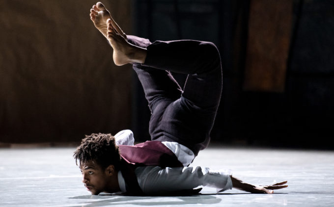 A dancer in a maroon vest and white shirt performs a floor move with his legs raised and crossed overhead.