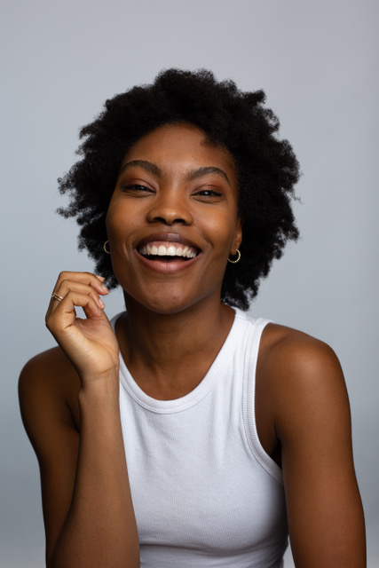 A person with a broad smile, wearing a white sleeveless top, holds a hand near their face. Their hair is styled in natural curls. The background is plain gray.