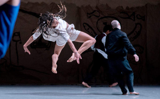 A dancer in mid-air with braided hair performs on stage, dressed in a white outfit, while two other dancers on the ground are partially visible.