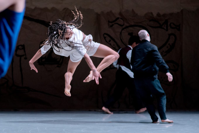 A dancer in mid-air with braided hair performs on stage, dressed in a white outfit, while two other dancers on the ground are partially visible.