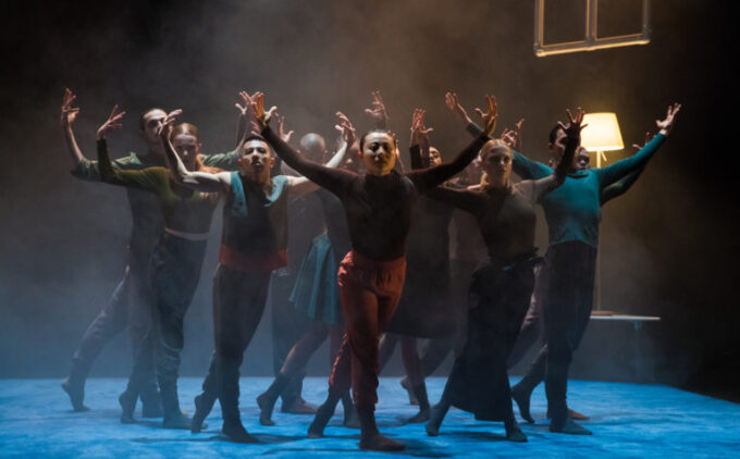 A group of dancers performs on a dimly lit stage with a blue floor. They pose with outstretched arms. A lamp and suspended frame are visible in the background.