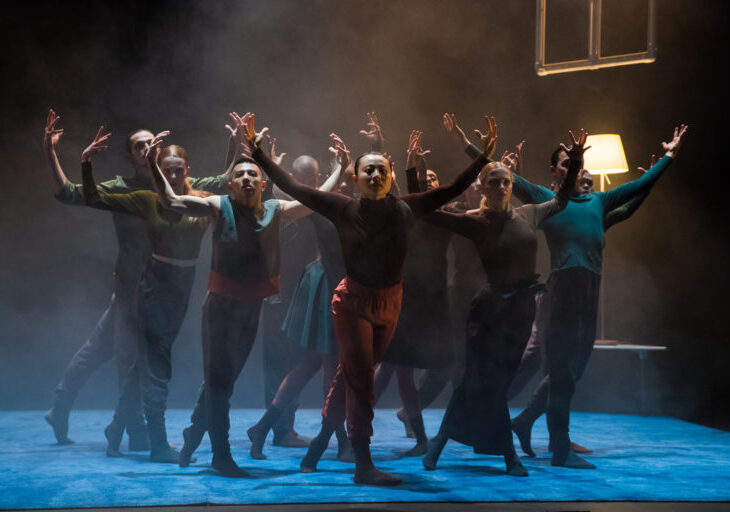 A group of dancers performs on a dimly lit stage with a blue floor. They pose with outstretched arms. A lamp and suspended frame are visible in the background.