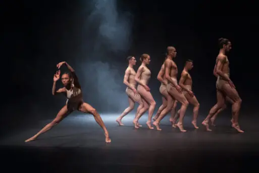 Group of six dancers in beige outfits performing on a dark stage, one dancer poses in the foreground while the others walk in a line in the background.