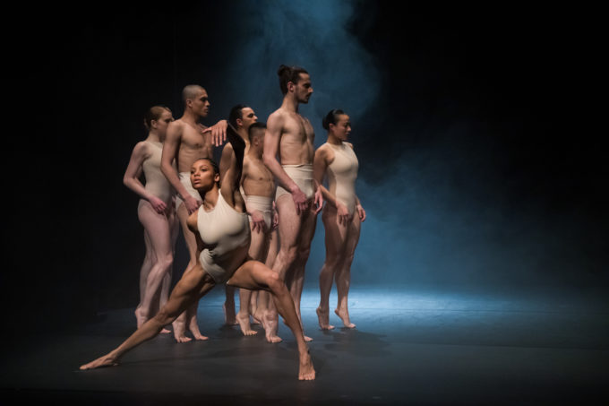 A group of six dancers in skin-tone costumes perform on a fog-covered stage under dramatic lighting.