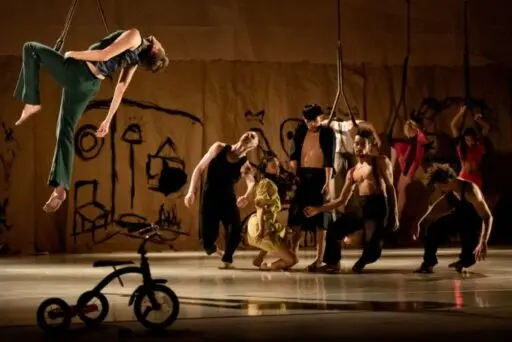 Performers in various poses on a stage with a drawn backdrop and a tricycle in the foreground. One person is suspended above the ground.