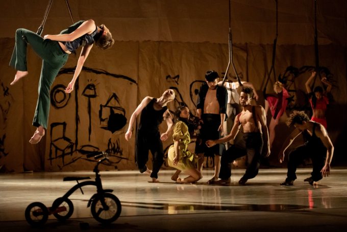 Performers in various poses on a stage with a drawn backdrop and a tricycle in the foreground. One person is suspended above the ground.