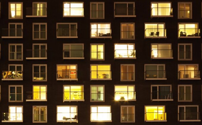 A nighttime view of a multi-story apartment building with lit windows showing various home interiors, some with visible furniture and decorations.