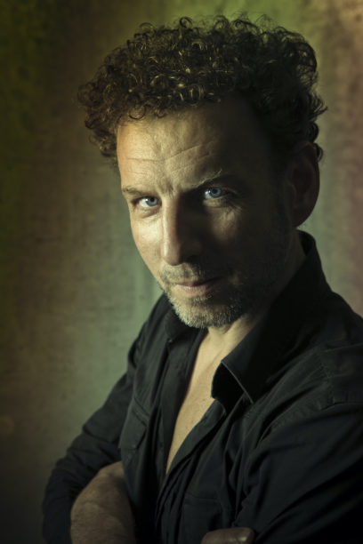 A man with curly hair and a beard looks directly at the camera, arms crossed, wearing a black shirt. The background is softly lit with subtle colors.