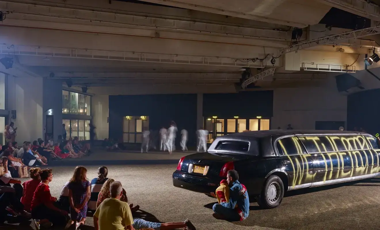 An audience looking at performers moving near a limousine. The black limousine has "We The People" in yellow spray paint.