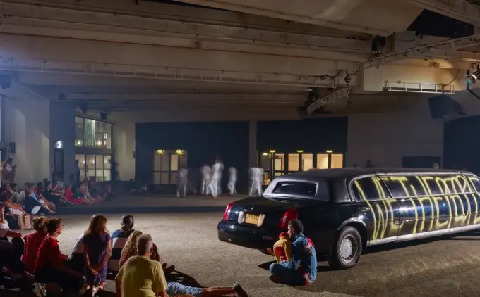 An audience looking at performers moving near a limousine. The black limousine has "We The People" in yellow spray paint.