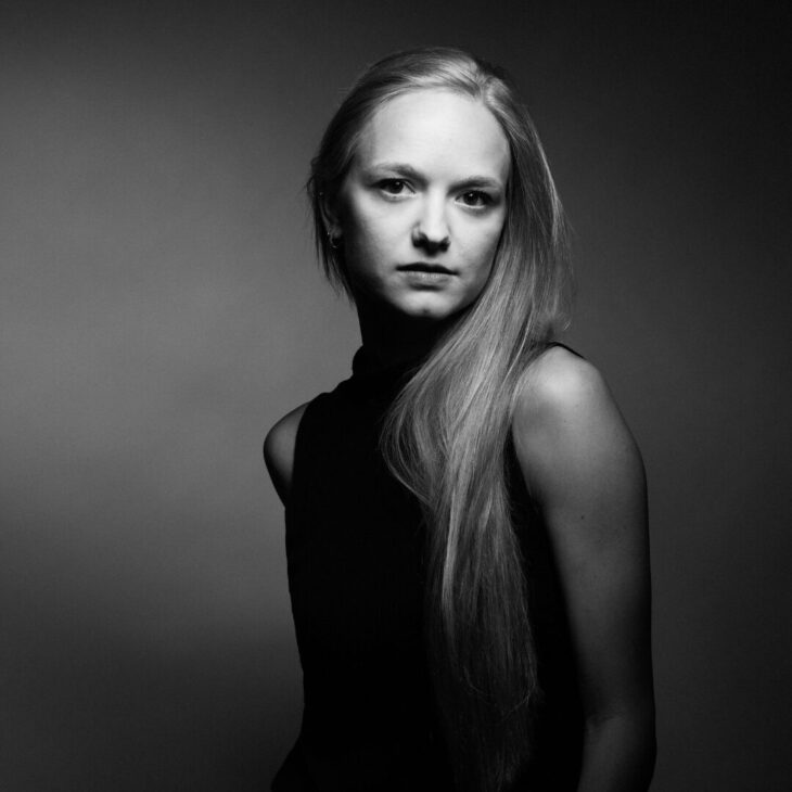 A black and white portrait of a person with long light hair, wearing a black short sleeve shirt, set against a plain background.