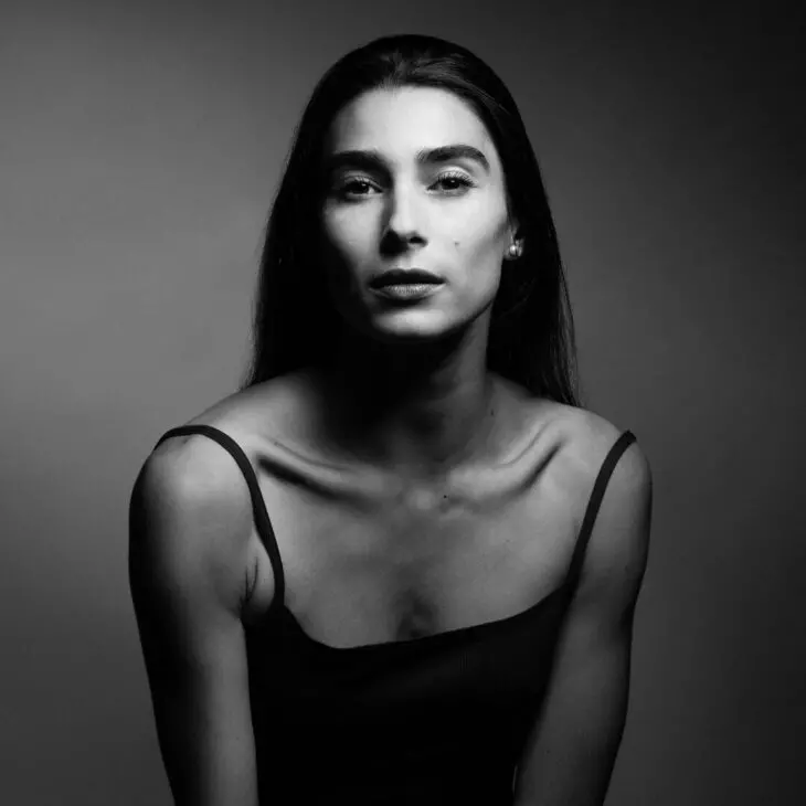 A black and white portrait of a person with long dark hair, wearing a black tank top, set against a plain background.